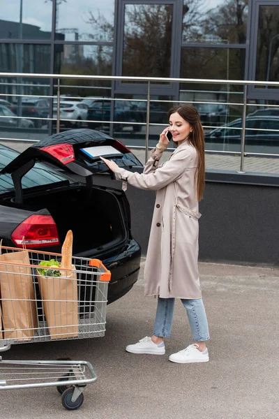 Young woman talking on cellphone near car trunk and shopping trolley outdoors — Stock Photo