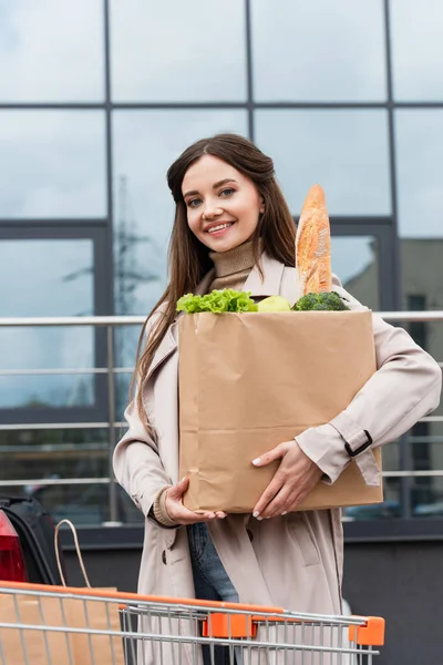 Mujer feliz mirando a la cámara mientras sostiene la bolsa de compras con alimentos frescos al aire libre - foto de stock