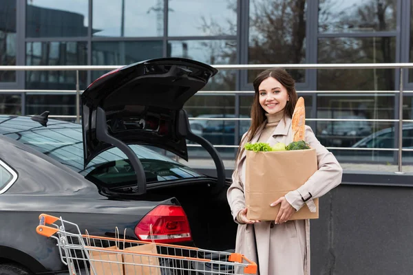 Femme joyeuse avec sac à provisions près du chariot et de la voiture avec coffre ouvert — Photo de stock