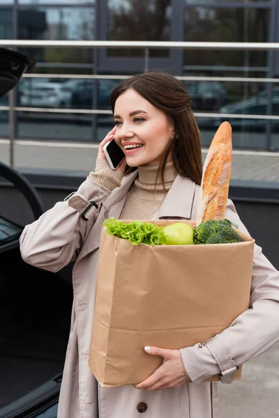 Donna sorridente che tiene la borsa della spesa mentre parla sullo smartphone all'aperto — Foto stock