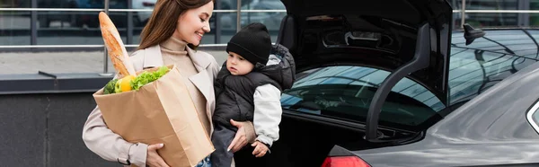 Усміхнена жінка з сином і сумки з покупками біля машини, банер — стокове фото