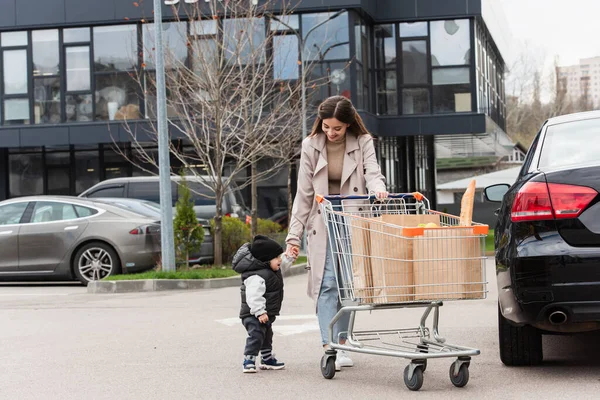 Mãe com filho criança andando no estacionamento do carro perto do carrinho de compras — Fotografia de Stock