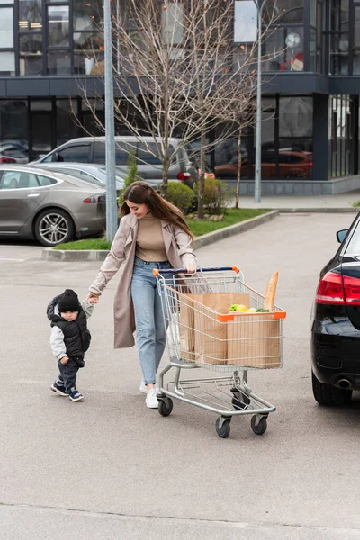 Молодая женщина, держась за руку ребенка во время прогулки с корзиной на автостоянке — стоковое фото