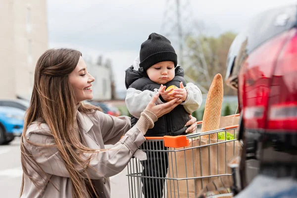 Smiling mother giving orange to toddler son sitting in shopping cart — Stock Photo