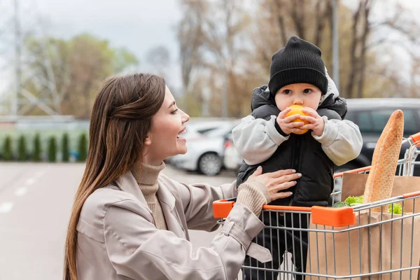 Bebé niño en la cesta de la compra sosteniendo naranja fresca cerca de madre feliz - foto de stock