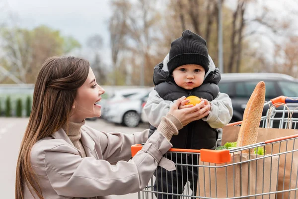 Donna felice guardando il bambino in possesso di arancia fresca mentre seduto nel carrello della spesa — Foto stock