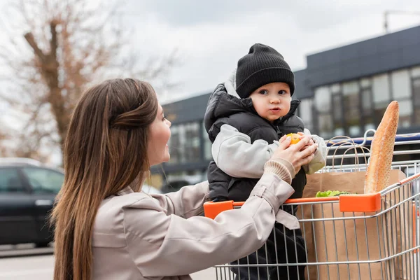 Bebé niño sosteniendo naranja fresca mientras está sentado en el carrito de compras con compras cerca de la madre - foto de stock