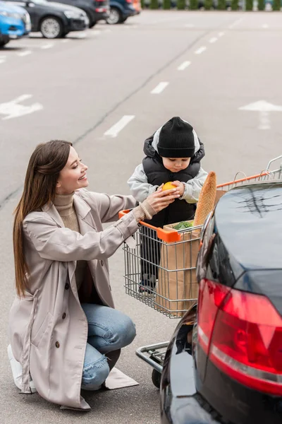 Femme souriante donnant orange à tout-petit enfant assis dans le panier — Photo de stock