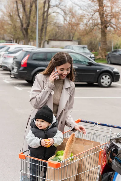 Mujer joven hablando en el teléfono inteligente cerca de compras y niño pequeño hijo en el carrito de la compra - foto de stock