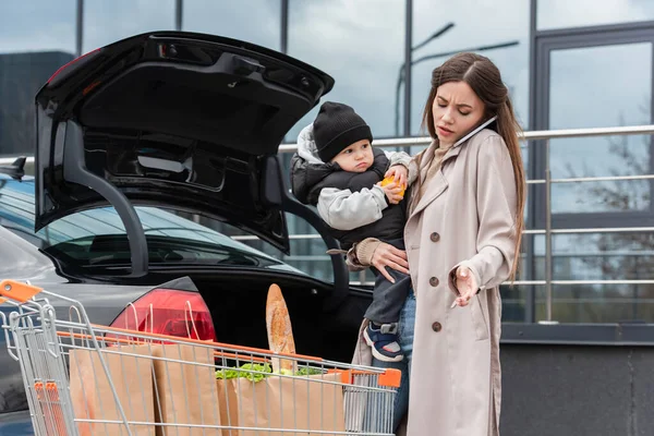 Молода жінка з сином в руках говорить на смартфоні біля машини і кошика для покупок — Stock Photo
