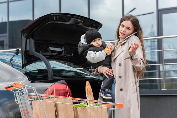 Woman talking on mobile phone while holding son near car and shopping cart — Stock Photo