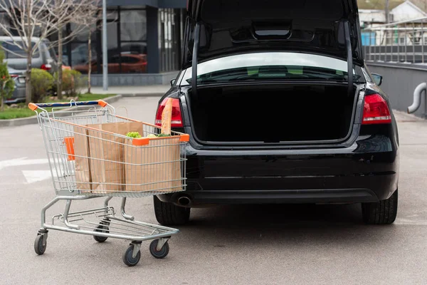 Shopping cart with fresh grocery near black car with open trunk — Stock Photo