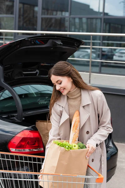 Jolie, souriante femme tenant sac à provisions près du coffre de voiture ouverte — Photo de stock