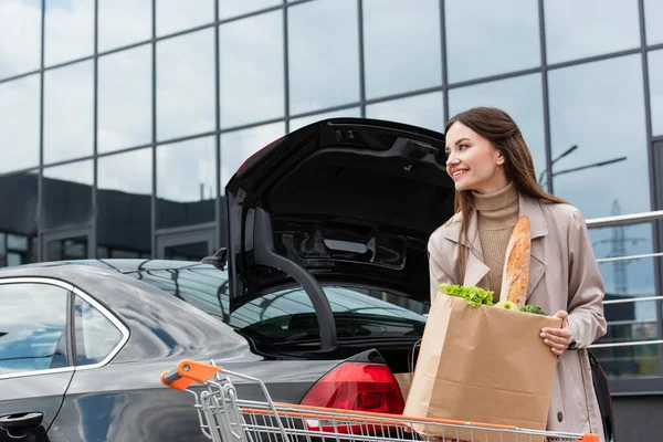Mujer feliz sosteniendo bolsa de compras cerca del coche con tronco abierto — Stock Photo