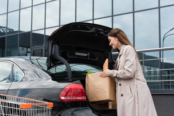 Young housewife putting shopping bag into car trunk on parking — Stock Photo