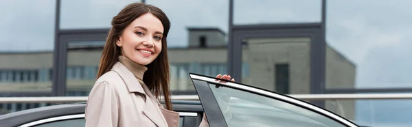 Pleased woman smiling at camera while standing near car, banner — Stock Photo