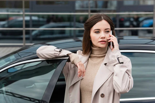 Pretty woman in trench coat calling on mobile phone near car — Stock Photo