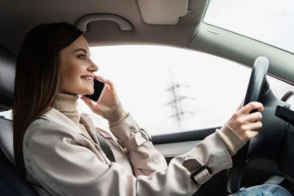 Vue latérale de la femme souriante conduisant une voiture et parlant sur smartphone — Photo de stock