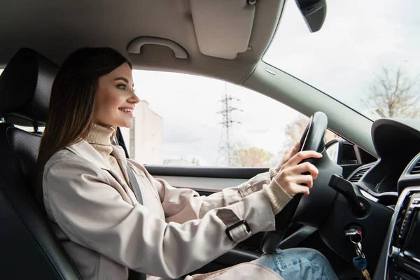 Side view of smiling woman in trench coat driving car in city — Stock Photo