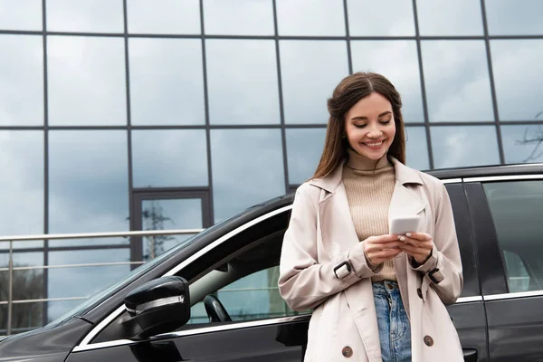 Fröhliche Frau im Trenchcoat nutzt Smartphone, während sie neben Auto steht — Stockfoto