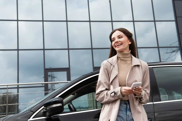 Fröhliche Frau mit Smartphone schaut weg, während sie neben Auto steht — Stockfoto