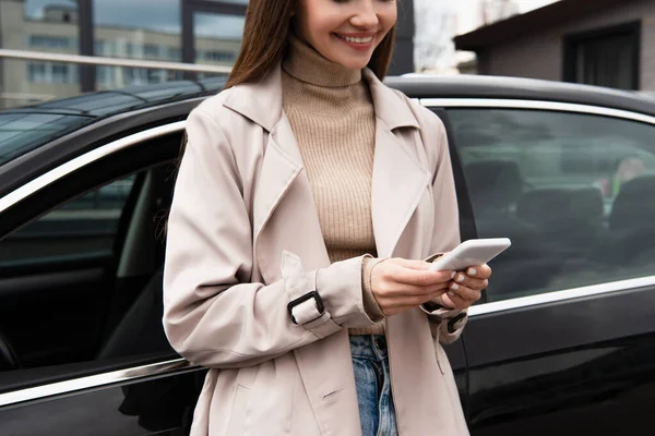 Vista ritagliata di donna felice in trench messaggistica sul telefono cellulare vicino all'auto — Foto stock