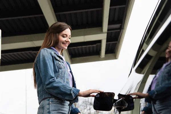 Vista basso angolo di donna sorridente in giacca di jeans in piedi vicino auto all'aperto — Foto stock