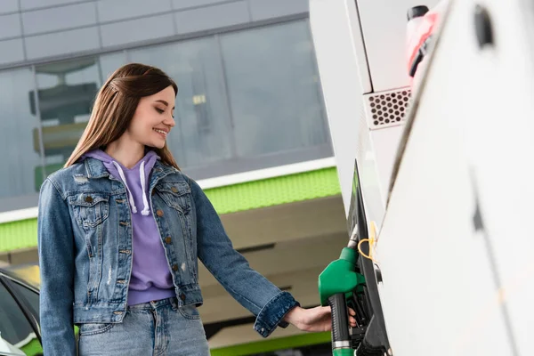 Mulher sorridente em jaqueta de ganga levando pistola de combustível no posto de gasolina — Fotografia de Stock