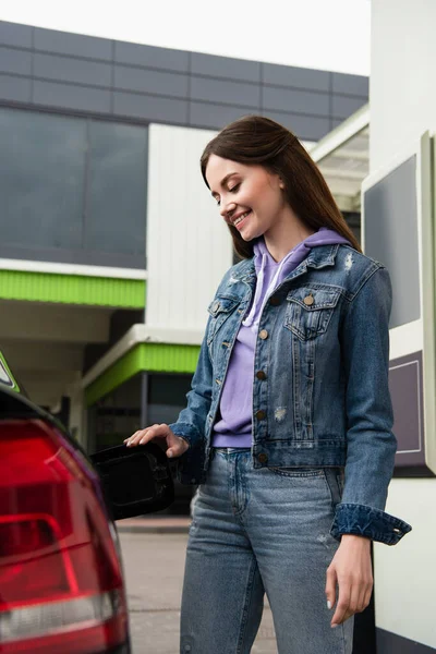 Happy woman in denim clothes opening fuel tank of car outdoors — Stock Photo