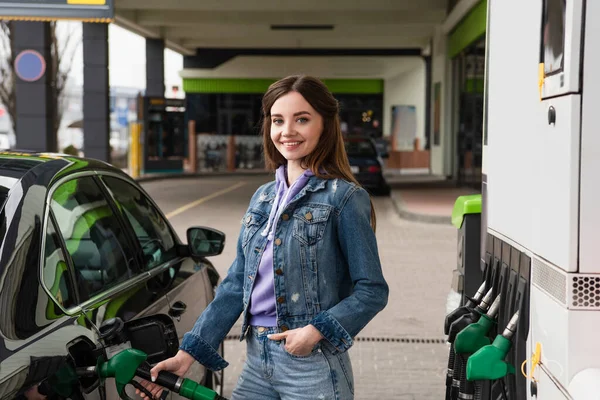 Jolie femme avec la main dans la poche de jeans voiture de remplissage sur la station-service — Photo de stock