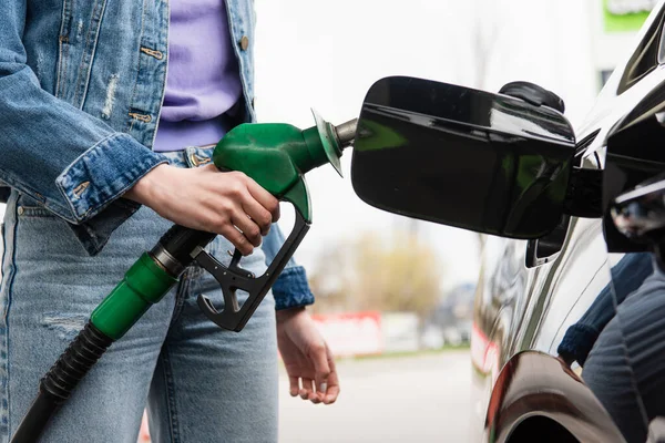 Visão parcial da jovem mulher em jeans segurando pistola de gasolina perto do automóvel — Fotografia de Stock