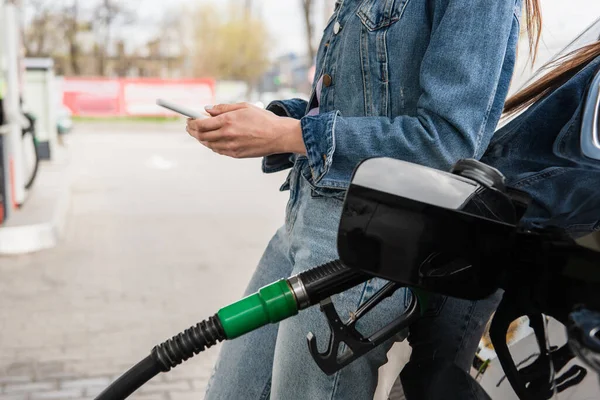 Teilbild einer jungen Frau, die auf Handy in der Nähe von Auto an Tankstelle Nachrichten sendet — Stockfoto