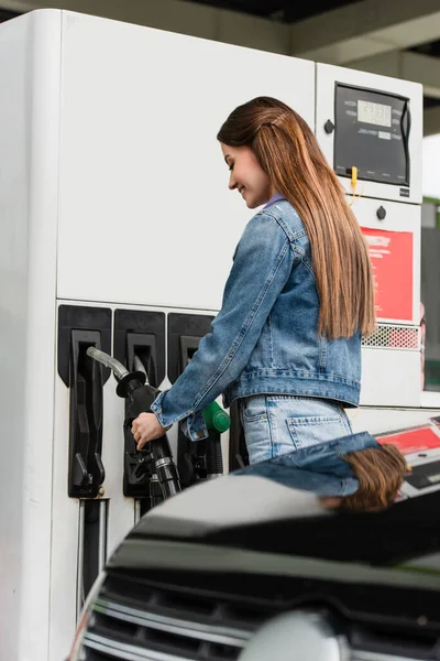 Femme souriante avec pistolet à carburant près de la voiture floue sur la gare routière — Photo de stock