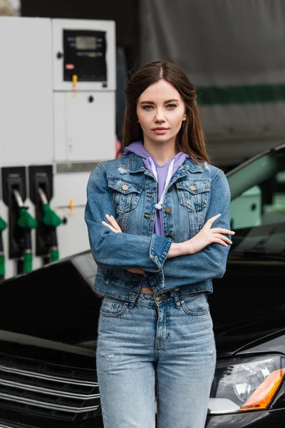 Giovane donna in abiti di denim in piedi con le braccia incrociate vicino auto al distributore di benzina — Foto stock