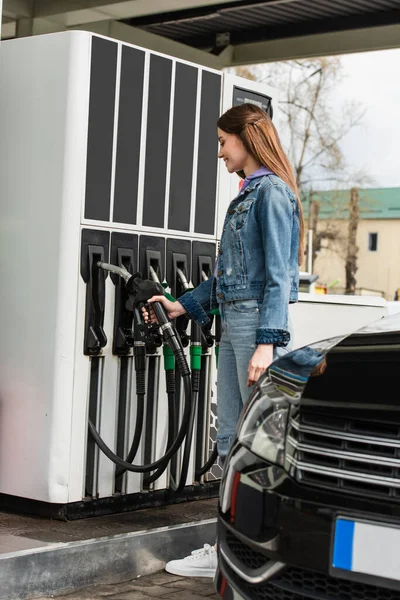 Joven mujer sosteniendo pistola de gasolina cerca de coche en la gasolinera - foto de stock