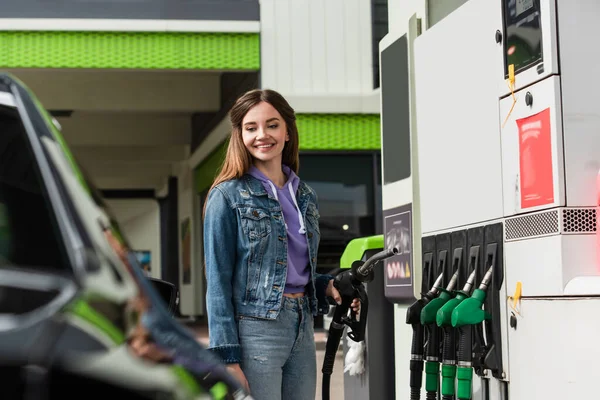 Femme souriante en vêtements denim tenant pistolet à essence près de voiture floue — Photo de stock