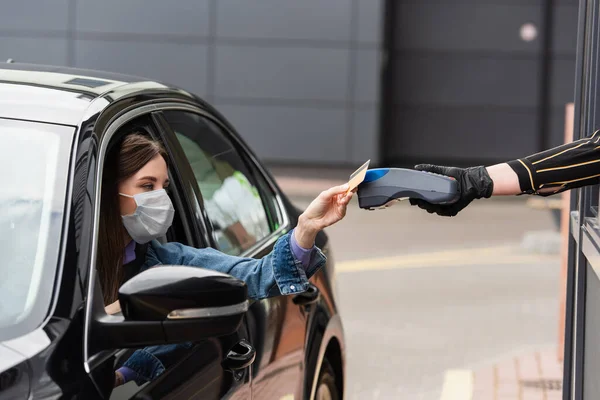 Mujer en máscara protectora que paga con tarjeta de crédito a través de la terminal mientras está sentado en el coche - foto de stock