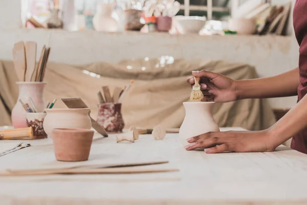 Partial view of young african american woman glazing clay pot with brush in pottery — Stock Photo