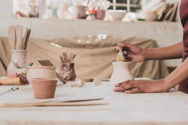 Vue partielle de la jeune femme afro-américaine glaçure pot d'argile avec brosse en poterie — Stock Photo