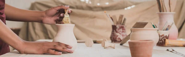 Частичный вид молодой африканской американки, остекляющей глиняный горшок кистью в керамике, баннер — стоковое фото