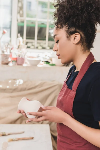 Serious young african american woman looking at clay pot in hands in pottery — Stock Photo