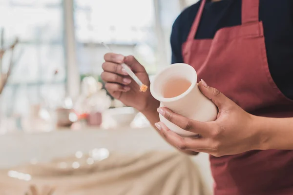 Partial view of young african american woman holding clay pot and brush in pottery — Stock Photo