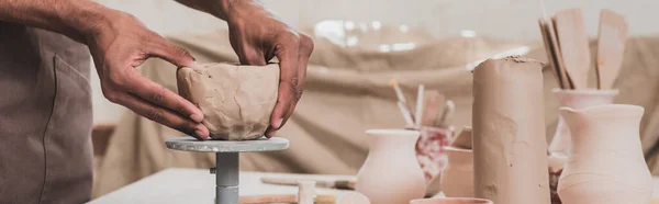 Vista parziale del giovane uomo afroamericano che scolpisce il vaso di argilla con le mani sul tavolo con attrezzature in ceramica, banner — Foto stock