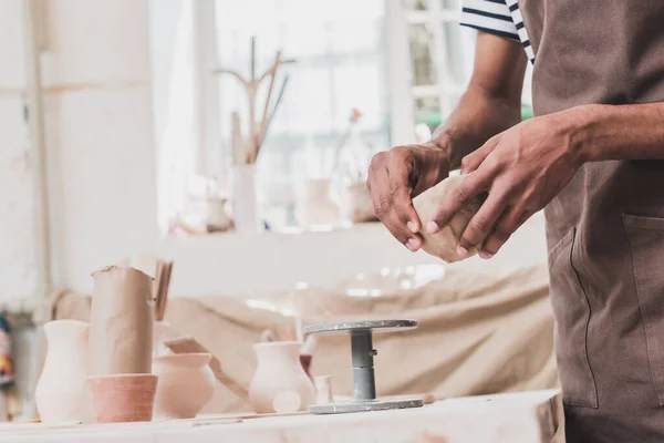 Vista parziale del giovane uomo afro-americano scolpire vaso di argilla con le mani vicino al tavolo con attrezzature in ceramica — Foto stock