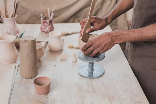 Vista parziale di giovane uomo afro-americano scolpire vaso di argilla con spatola vicino al tavolo con attrezzature in ceramica — Foto stock