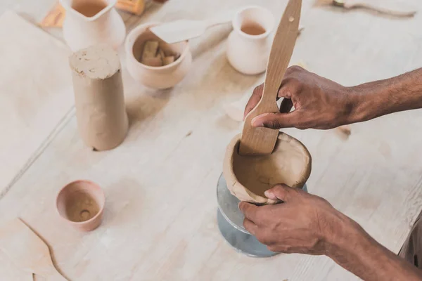 Partial view of young african american man sculpting clay pot with spatula on table with equipment in pottery — Stock Photo