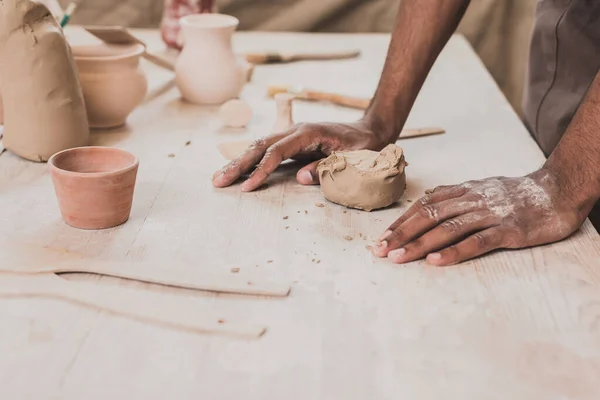 Partial view of young african american man with hands near piece of clay on table with equipment in pottery — Stock Photo