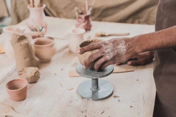 Teilansicht eines jungen afrikanisch-amerikanischen Mannes, der Tongefäße mit der Hand auf einem Tisch mit Geräten aus Keramik gestaltet — Stockfoto