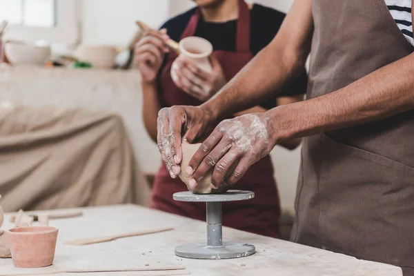 Vista parziale del giovane uomo afro-americano scolpire vaso di argilla con le mani vicino alla donna sfocata in ceramica — Foto stock