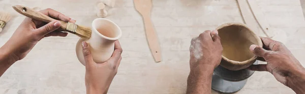 Partial view of young african american couple sculpting clay pots with hands and brush in pottery, banner — Stock Photo